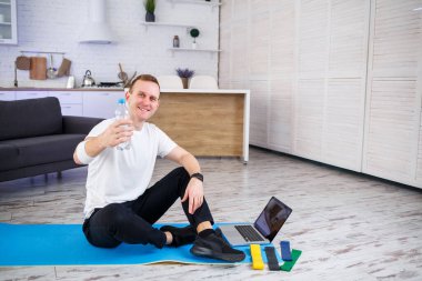 Yakışıklı bir genç adam yerde oturmuş, elinde bir şişe su tutarken internetten spor yapıyor.