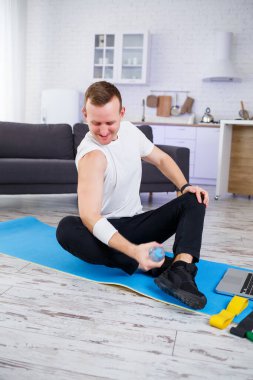 Yakışıklı bir genç adam yerde oturmuş, elinde bir şişe su tutarken internetten spor yapıyor.