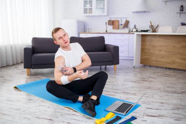 Yakışıklı bir genç adam yerde oturmuş, elinde bir şişe su tutarken internetten spor yapıyor.