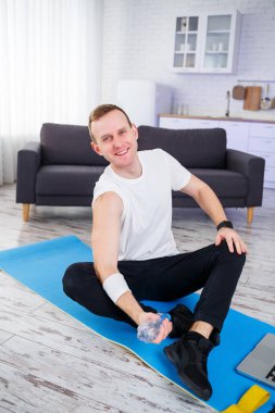 Yakışıklı bir genç adam yerde oturmuş, elinde bir şişe su tutarken internetten spor yapıyor.