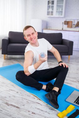 Yakışıklı bir genç adam yerde oturmuş, elinde bir şişe su tutarken internetten spor yapıyor.