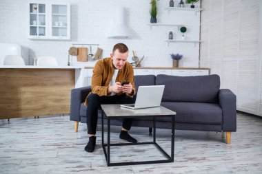 Başarılı genç adam evinde, kanepede dizüstü bilgisayarıyla oturuyor ve çalışıyor. Karantina sırasında uzak çalışma.