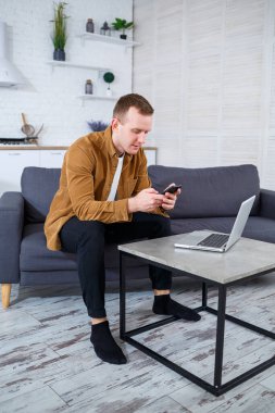 Başarılı genç adam evinde, kanepede dizüstü bilgisayarıyla oturuyor ve çalışıyor. Karantina sırasında uzak çalışma.