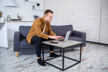 Başarılı genç adam evinde, kanepede dizüstü bilgisayarıyla oturuyor ve çalışıyor. Karantina sırasında uzak çalışma.