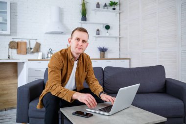 Başarılı genç adam evinde, kanepede dizüstü bilgisayarıyla oturuyor ve çalışıyor. Karantina sırasında uzak çalışma.