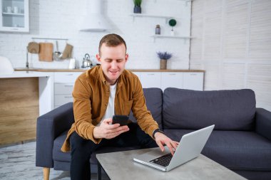 Başarılı genç adam evinde, kanepede dizüstü bilgisayarıyla oturuyor ve çalışıyor. Karantina sırasında uzak çalışma.