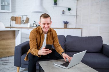 Başarılı genç adam evinde, kanepede dizüstü bilgisayarıyla oturuyor ve çalışıyor. Karantina sırasında uzak çalışma.