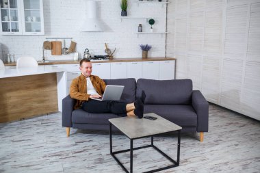 Başarılı genç bir adam evinde, kanepede dizüstü bilgisayarıyla oturuyor ve çalışıyor. Karantina sırasında uzak çalışma.