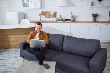 Başarılı genç bir adam evinde, kanepede dizüstü bilgisayarıyla oturuyor ve çalışıyor. Karantina sırasında uzak çalışma.