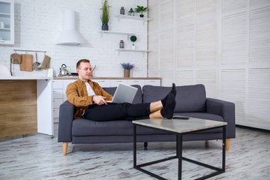 Başarılı genç bir adam evinde, kanepede dizüstü bilgisayarıyla oturuyor ve çalışıyor. Karantina sırasında uzak çalışma.