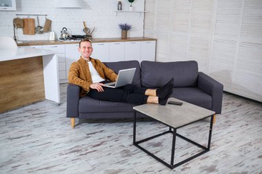 Başarılı genç bir adam evinde, kanepede dizüstü bilgisayarıyla oturuyor ve çalışıyor. Karantina sırasında uzak çalışma.