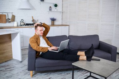 Başarılı genç bir adam evinde, kanepede dizüstü bilgisayarıyla oturuyor ve çalışıyor. Karantina sırasında uzak çalışma.