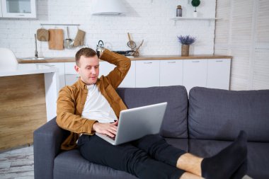 Başarılı genç bir adam evinde, kanepede dizüstü bilgisayarıyla oturuyor ve çalışıyor. Karantina sırasında uzak çalışma.