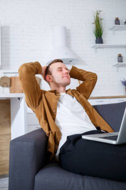 Başarılı genç bir adam evinde, kanepede dizüstü bilgisayarıyla oturuyor ve çalışıyor. Karantina sırasında uzak çalışma.