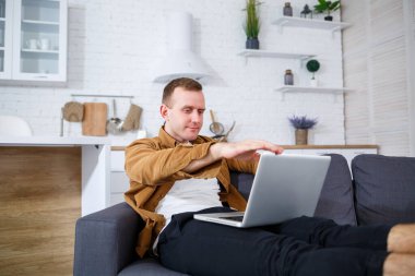 Başarılı genç bir adam evinde, kanepede dizüstü bilgisayarıyla oturuyor ve çalışıyor. Karantina sırasında uzak çalışma.