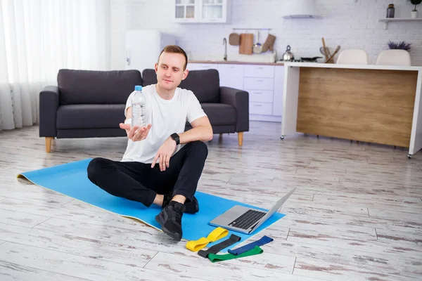 The man goes in for sports at home. Athlete drinks water while sitting on the floor at home, healthy lifestyle