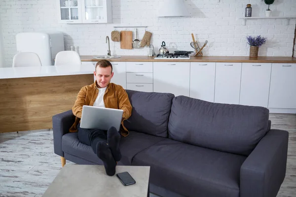 Başarılı genç bir adam evinde, kanepede dizüstü bilgisayarıyla oturuyor ve çalışıyor. Karantina sırasında uzak çalışma.