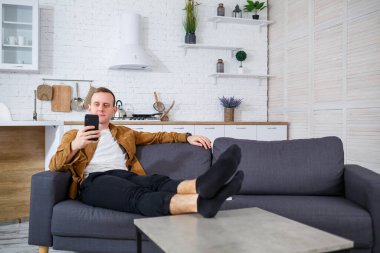 Genç ve başarılı bir adam evinde kanepede oturuyor elinde telefon ve video bağlantısı ile konuşuyor. Karantina sırasında uzak çalışma.