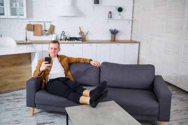 Genç ve başarılı bir adam evinde kanepede oturuyor elinde telefon ve video bağlantısı ile konuşuyor. Karantina sırasında uzak çalışma.