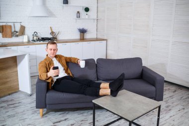 Genç ve başarılı bir adam evinde kanepede oturuyor elinde telefon ve video bağlantısı ile konuşuyor. Karantina sırasında uzak çalışma.