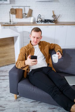 Genç ve başarılı bir adam evinde kanepede oturuyor elinde telefon ve video bağlantısı ile konuşuyor. Karantina sırasında uzak çalışma.