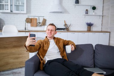 Genç ve başarılı bir adam evinde kanepede oturuyor elinde telefon ve video bağlantısı ile konuşuyor. Karantina sırasında uzak çalışma.