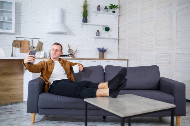 Genç ve başarılı bir adam evinde kanepede oturuyor elinde telefon ve video bağlantısı ile konuşuyor. Karantina sırasında uzak çalışma.