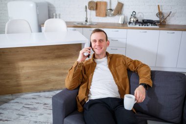 Mutlu genç adam oturma odasındaki kanepede oturuyor bir fincan kahveyle ve telefonla konuşuyor..