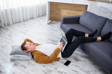 Çekici, güler yüzlü, gündelik giysili genç adam oturma odasındaki koltukta dizüstü bilgisayar kullanarak oturuyor.