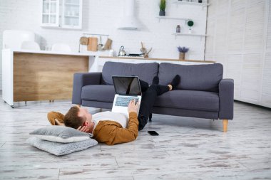 Çekici, güler yüzlü, gündelik giysili genç adam oturma odasındaki koltukta dizüstü bilgisayar kullanarak oturuyor.