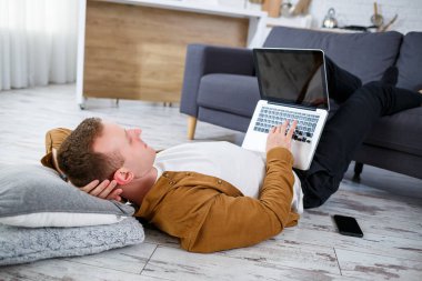 Çekici, güler yüzlü, gündelik giysili genç adam oturma odasındaki koltukta dizüstü bilgisayar kullanarak oturuyor.