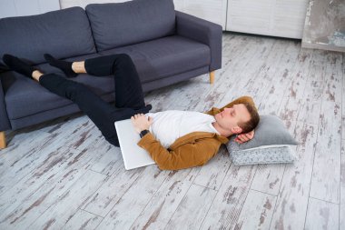 Çekici, güler yüzlü, gündelik giysili genç adam oturma odasındaki koltukta dizüstü bilgisayar kullanarak oturuyor.