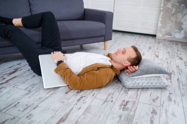 Çekici, güler yüzlü, gündelik giysili genç adam oturma odasındaki koltukta dizüstü bilgisayar kullanarak oturuyor.