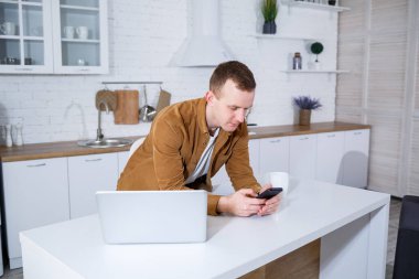 Genç bir adam mutfakta dizüstü bilgisayarla oturuyor ve telefonla konuşuyor. Uzak çalışma.