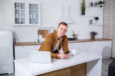 Genç bir adam mutfakta dizüstü bilgisayarla oturuyor ve telefonla konuşuyor. Uzak çalışma.