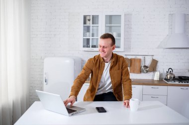 Genç bir adam mutfakta dizüstü bilgisayarla oturuyor ve telefonla konuşuyor. Uzak çalışma.