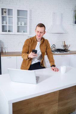 Genç bir adam mutfakta dizüstü bilgisayarla oturuyor ve telefonla konuşuyor. Uzak çalışma.