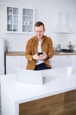 Genç bir adam mutfakta dizüstü bilgisayarla oturuyor ve telefonla konuşuyor. Uzak çalışma.