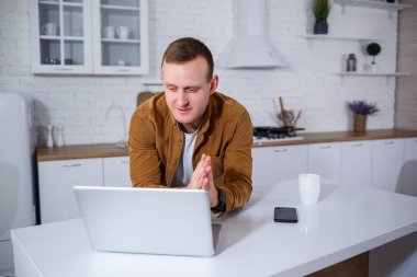 Gündelik giysiler içinde çekici bir genç adam mutfakta dizüstü bilgisayar kullanarak oturuyor. Evden çalışmak, uzak iş akışı.