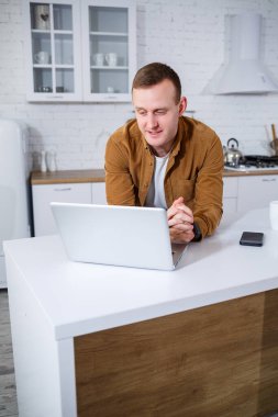 Gündelik giysiler içinde çekici bir genç adam mutfakta dizüstü bilgisayar kullanarak oturuyor. Evden çalışmak, uzak iş akışı.