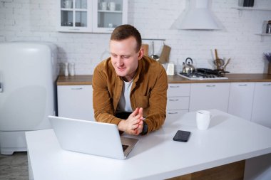 Gündelik giysiler içinde çekici bir genç adam mutfakta dizüstü bilgisayar kullanarak oturuyor. Evden çalışmak, uzak iş akışı.