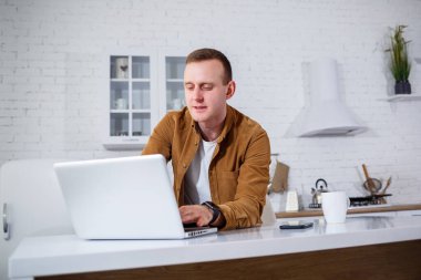 Gündelik giysiler içinde çekici bir genç adam mutfakta dizüstü bilgisayar kullanarak oturuyor. Evden çalışmak, uzak iş akışı.