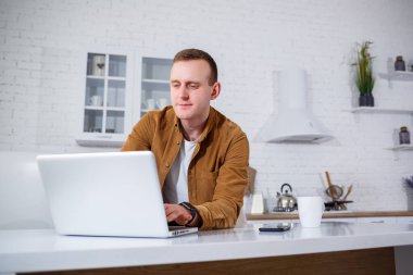Gündelik giysiler içinde çekici bir genç adam mutfakta dizüstü bilgisayar kullanarak oturuyor. Evden çalışmak, uzak iş akışı.
