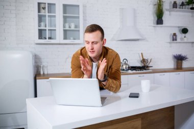 Gündelik giysiler içinde çekici bir genç adam mutfakta dizüstü bilgisayar kullanarak oturuyor. Evden çalışmak, uzak iş akışı.