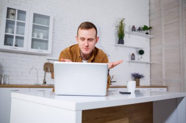 Gündelik giysiler içinde çekici bir genç adam mutfakta dizüstü bilgisayar kullanarak oturuyor. Evden çalışmak, uzak iş akışı.