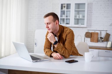Gündelik giysiler içinde çekici bir genç adam mutfakta dizüstü bilgisayar kullanarak oturuyor. Evden çalışmak, uzak iş akışı.