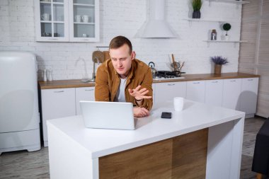 Gündelik giysiler içinde çekici bir genç adam mutfakta dizüstü bilgisayar kullanarak oturuyor. Evden çalışmak, uzak iş akışı.