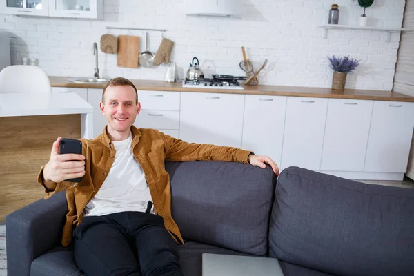 Genç ve başarılı bir adam evinde kanepede oturuyor elinde telefon ve video bağlantısı ile konuşuyor. Karantina sırasında uzak çalışma.