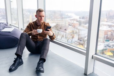 Yakışıklı genç erkek yönetici panoramik pencerenin yanında telefonla yerde oturuyor. Erkek işadamı yeni bir proje üzerinde çalışıyor.