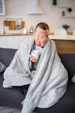 Genç adam battaniyeye sarılı bir fincan çayla kanepede oturuyor ve hasta.